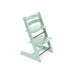 A pale green wooden high chair stands at an angle, showcasing its adjustable seat and footrest for growing children. 
