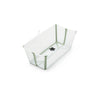 A clear plastic baby bathtub with sage green trim and a black drain plug sits at an angle, ready for bath time. 
